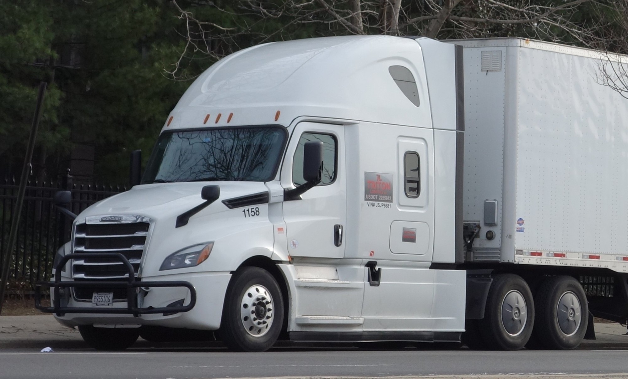 White American Freightliner tractor with an enclosed box trailer parked beside trees on a Queens street.