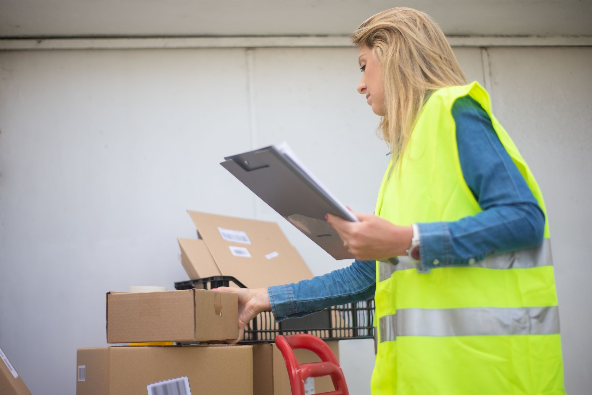 Warehouse worker in a high visibility vest holding a clipboard beside stacked cartons.